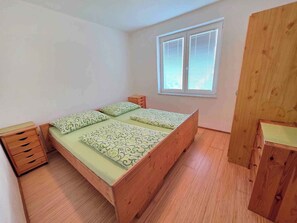 Et lille, rent værelse med en træseng, et natbord, et vindue med skodder og en træskab.