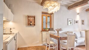 A kitchen with white cabinets, a wooden dining table, and chairs. There is a window with a view of the outside, and a radiator below the window. - Appartamenti Anguria Toscana One-bedroom (Sinalunga)