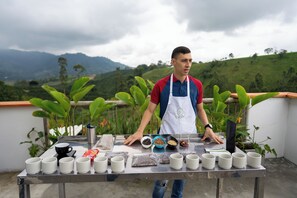 Una persona de pie detrás de una mesa con diversos artículos, incluyendo tazas, cuencos e individuales, frente a un fondo de colinas verdes y un cielo nublado.