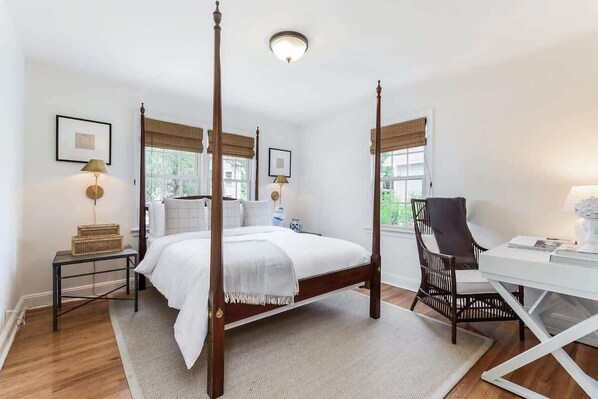 Una cama con dosel con cubrecama blanco, una mesita de noche de madera con lámpara, una silla de mimbre y un escritorio blanco con lámpara.