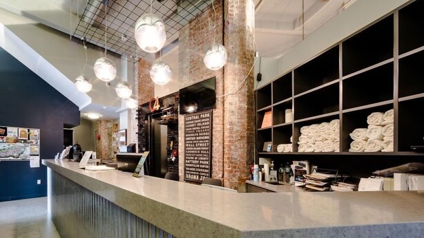 A hotel reception area with a modern design, featuring a counter with a digital display, a brick accent wall, and a large window with neatly folded towels. - Q4 Hotel & Hostel (Long Island City)