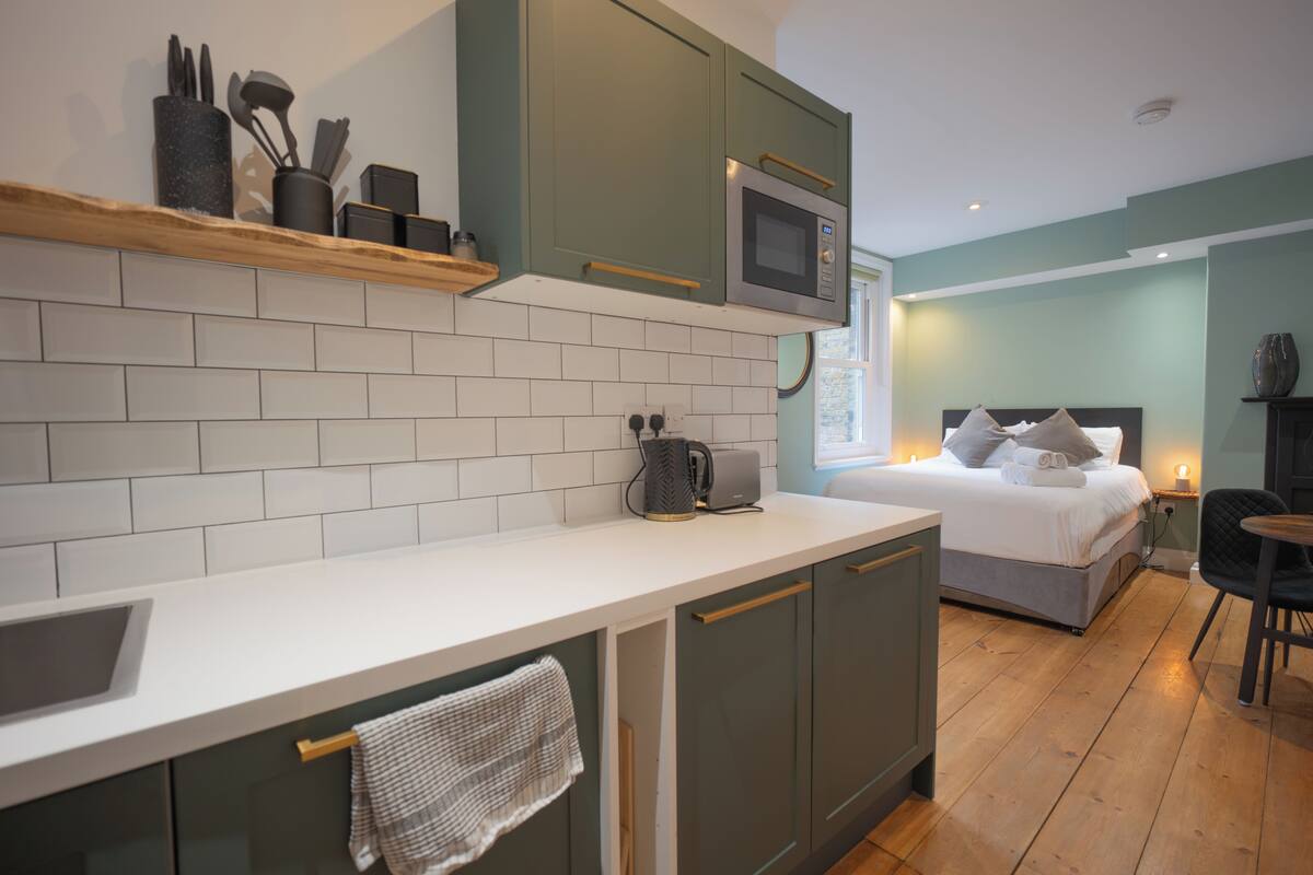 A compact kitchen with a microwave, a kettle, and a coffee maker on the counter. There is a bed with pillows and a bedside table with a lamp.