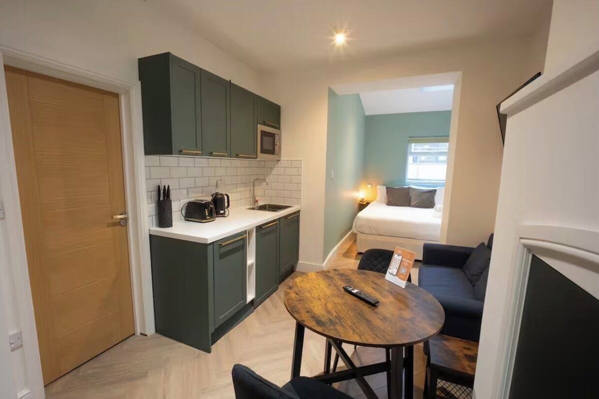 A compact kitchen with dark green cabinets, a microwave, and a coffee maker. There is a small dining table with a remote control. The room has a bed with a blue wall and a window.