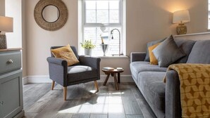 Un salon avec un canapé gris, un fauteuil bleu, une petite table d’appoint, un miroir décoratif et une fenêtre avec des stores.