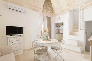 Una sala de estar moderna con televisión, muebles blancos, una mesa de comedor y una cocina al fondo.