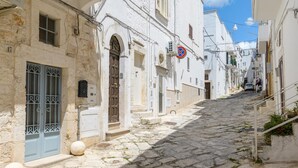 A narrow cobblestone street flanked by white buildings with arched doorways and barred windows. - In Alto Mare by Wonderful Italy (Ostuni)