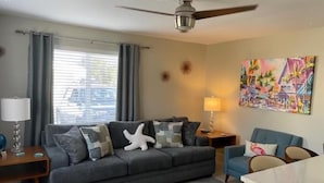 A living room with a grey sofa, a blue armchair, a dining table, a ceiling fan, and a colorful painting on the wall. - The Turtle Nest 6 in Fort Myers Beach (Fort Myers Beach)