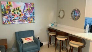 A living room with a blue armchair, a colorful painting, and a kitchenette with bar stools. - The Turtle Nest 6 in Fort Myers Beach (Fort Myers Beach)