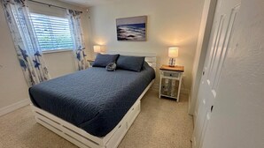 Une chambre à coucher avec un lit, deux tables de chevet, une commode, un tableau et une fenêtre avec des rideaux.