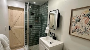 A bathroom with a shower, a sink, a wooden door, and a framed floral artwork on the wall.