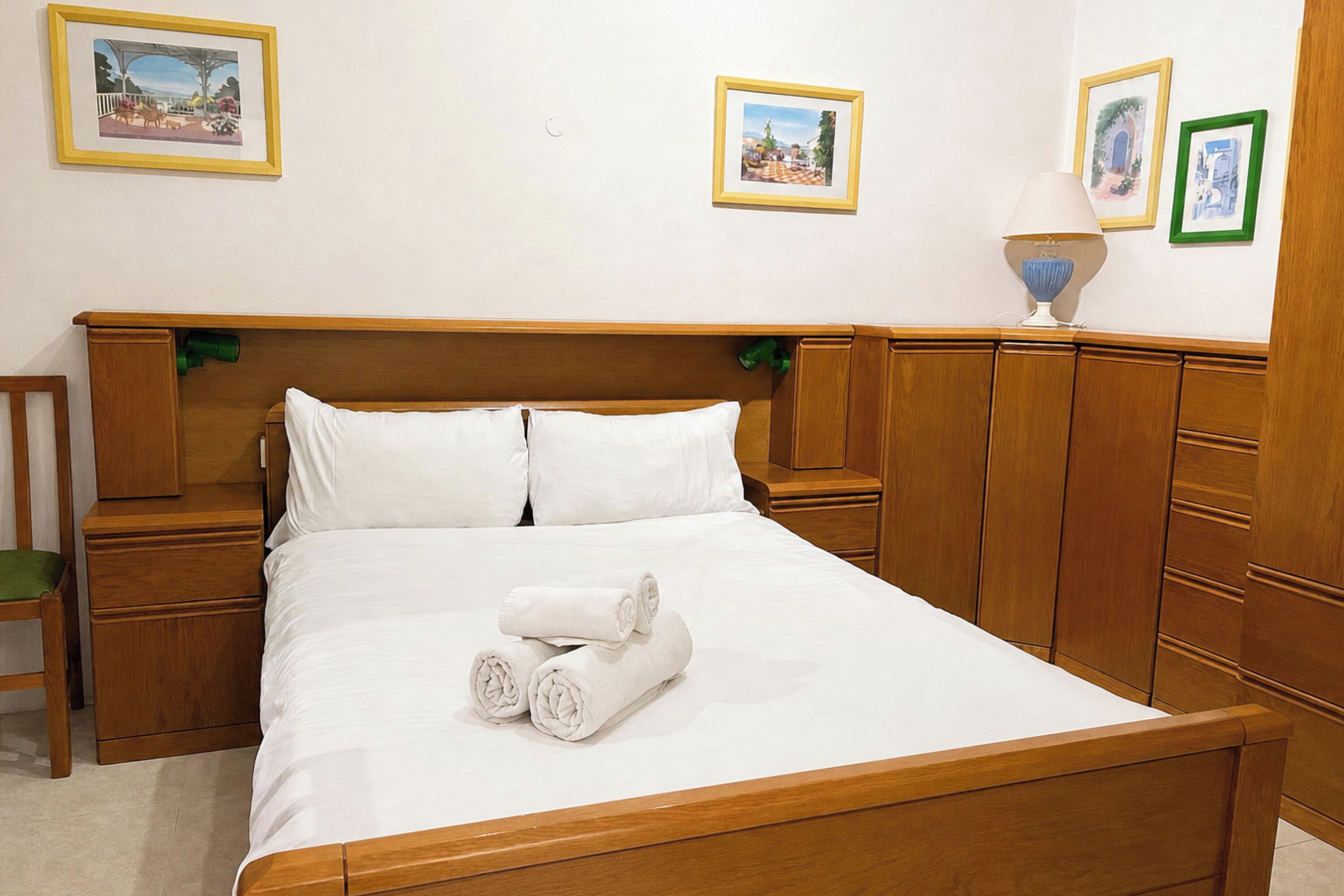 Una cama bien hecha con sábanas blancas y toallas dobladas, cabecera de madera, mesitas de noche de madera y una silla verde.