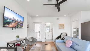 A modern living room with a cityscape TV, a grey armchair, a blue sofa, a wooden coffee table, and a ceiling fan. - East Downtown 3BR Condo Free Parking Sleeps 7 (Austin)
