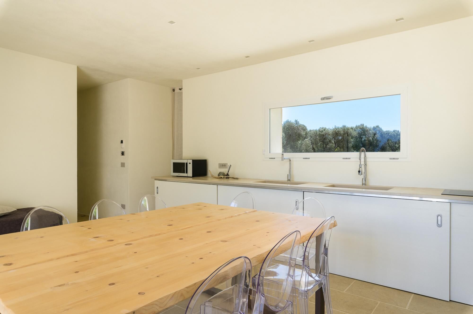 Una cocina moderna con una mesa de comedor de madera, sillas traslúcidas, microondas integrado y ventana con vistas a árboles.