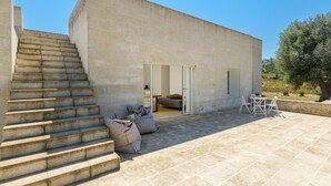 A modern building with a staircase leading to a door, a small outdoor seating area with a table and chairs, and a garden with trees. - Villa La Guaceta by Wonderful Italy (Carovigno)