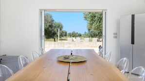 A modern dining area with a wooden table and clear chairs, a view of a garden and trees, and a white wall. - Villa La Guaceta by Wonderful Italy (Carovigno)
