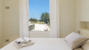 A hotel room with a bed, a window with white curtains, a view of the countryside, and a neatly folded towel on the bed. - Villa La Guaceta by Wonderful Italy (Carovigno)