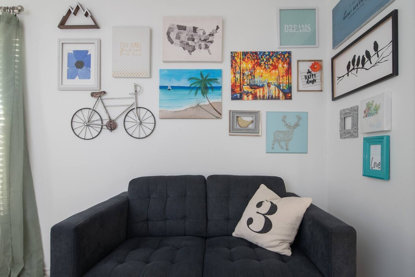 A living room with a dark grey sofa, a teal pillow, and various framed artworks on the wall, including a map, a beach scene, and a bird illustration.