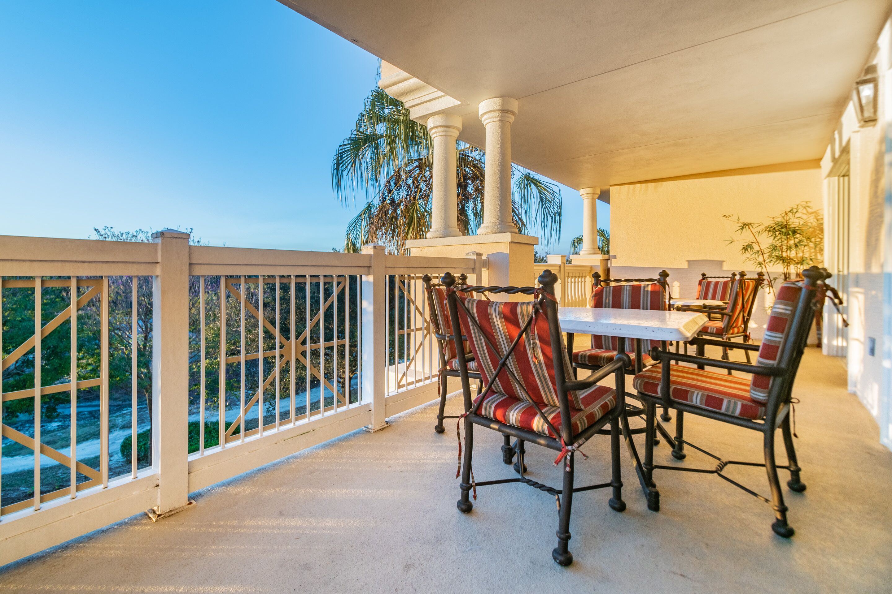 An outdoor patio with a table and chairs, a white railing, and palm trees in the background.