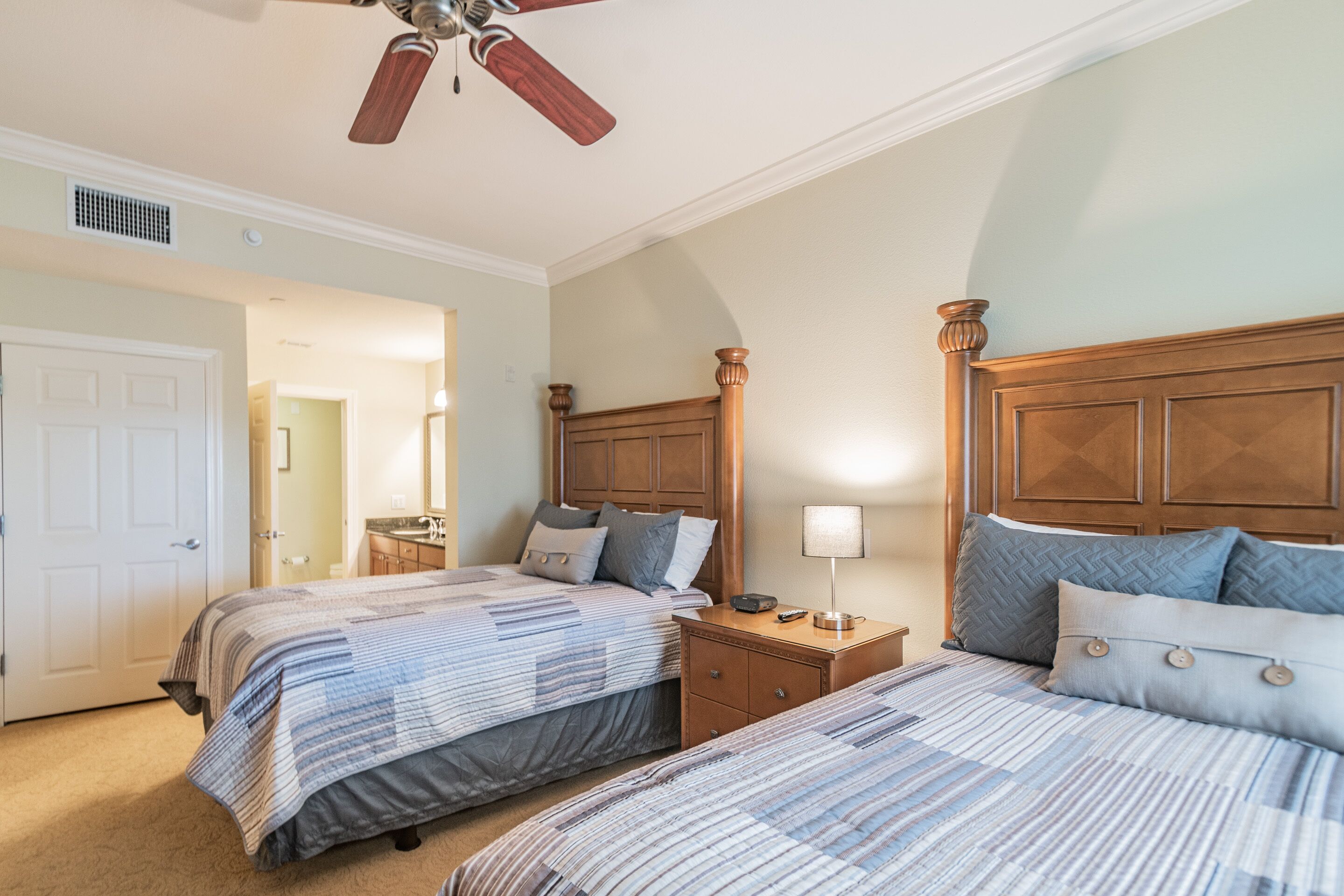 A bedroom with two beds, wooden headboards, a nightstand with a lamp, and a ceiling fan.