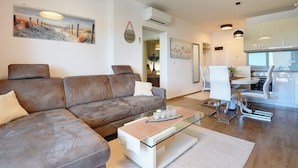 A modern living room with a brown leather sofa, a glass coffee table, and a dining area with white chairs. There is a painting on the wall and an air conditioning unit mounted above the sofa. - Modern apartment with sea view (Medulin)