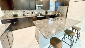 A modern kitchen with dark cabinets, granite countertops, stainless steel appliances, and a central island with stools. - Disney Dream House, Themed Rooms Pool Arcade BBQ! (Four Corners)