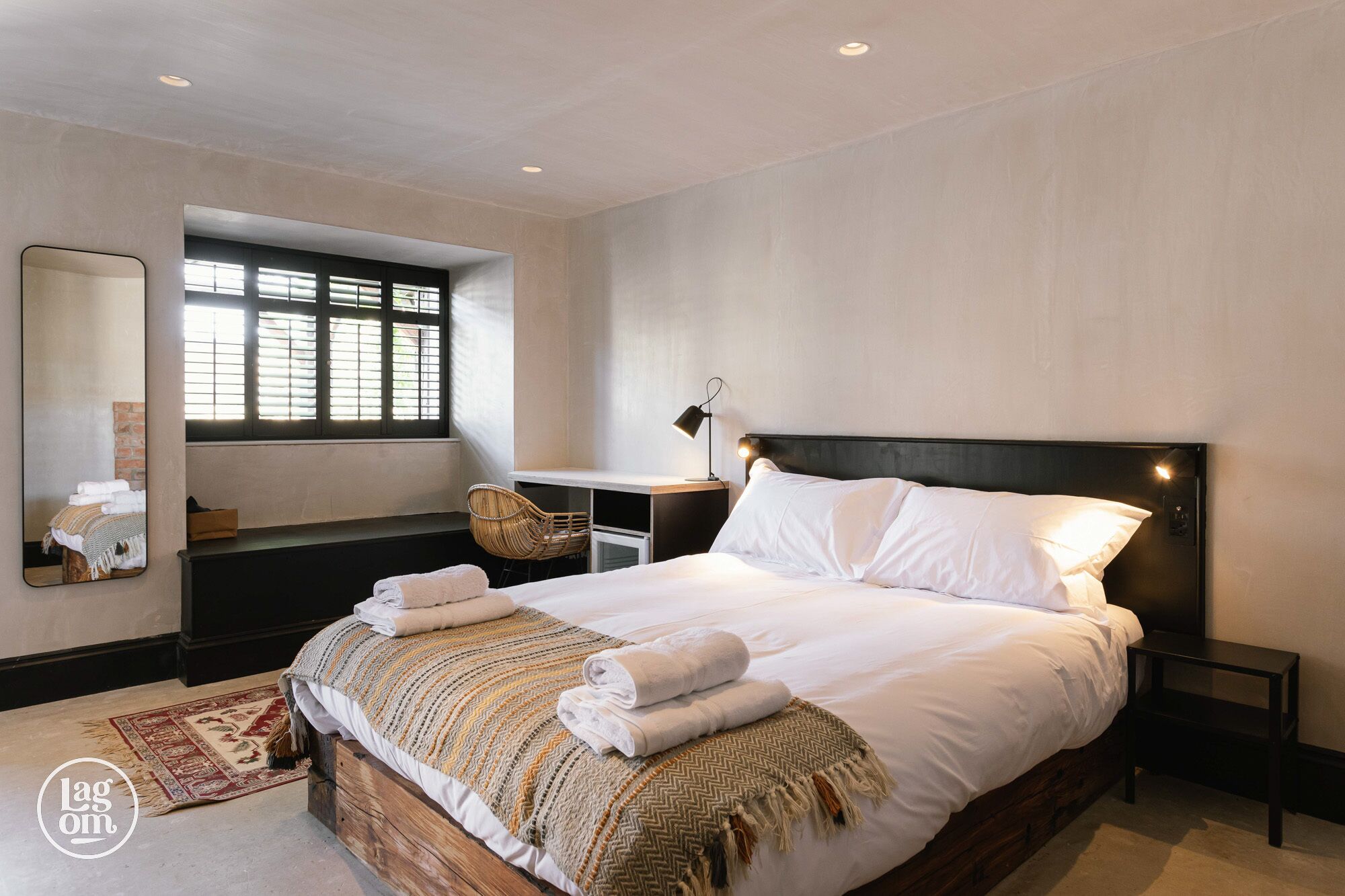 A modern bedroom with a large bed, a bedside table with a lamp, a chair, and a window with blinds.