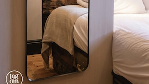 A bedroom with a stone wall, a bed with white linens, a wooden nightstand with a bottle and glasses, and a mirror reflecting the room. - Stay Lagom Lodge (Ballycastle)