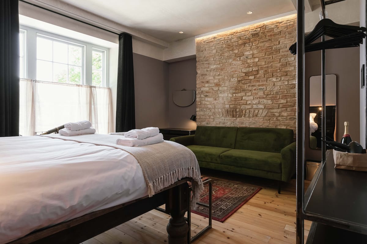 A neatly made bed with white linens and a green throw blanket, a green sofa, a brick accent wall, and a wooden floor.