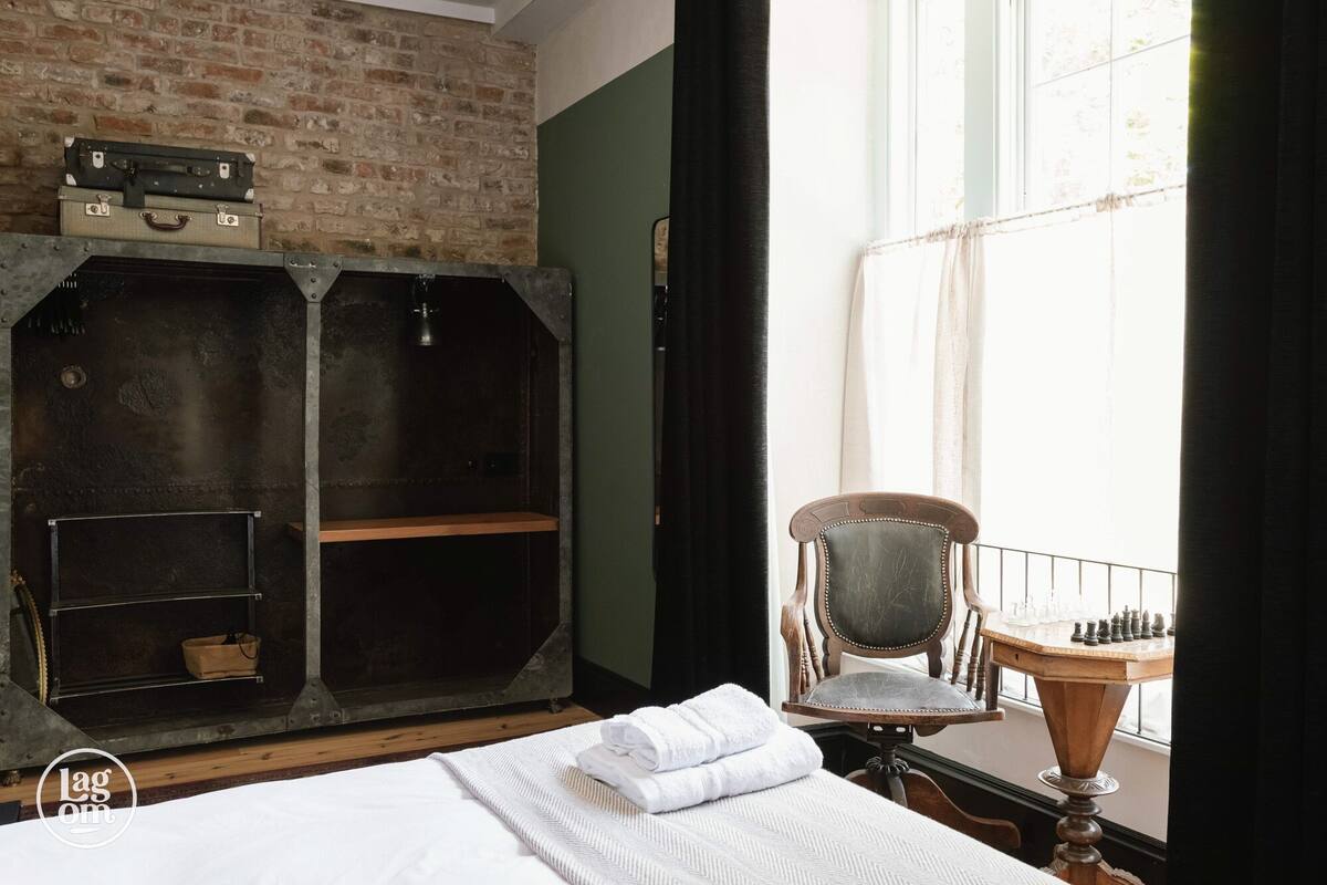 A room with a bed, a wooden chair, a chessboard, and a metal wardrobe with suitcases.