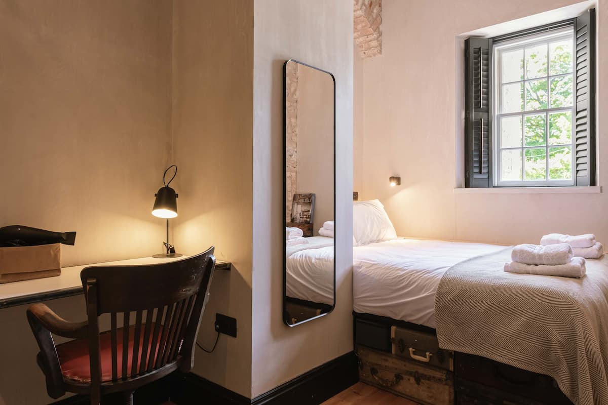 A bedroom with a bed, a desk with a chair, a mirror, and a window with shutters.
