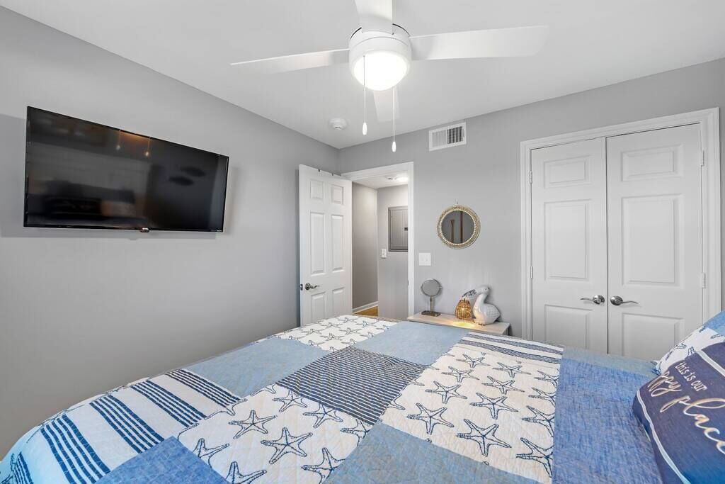 A bedroom with a bed, a ceiling fan, a TV mounted on the wall, and a door leading to another room.
