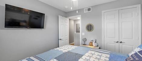 A bedroom with a bed, a ceiling fan, a TV mounted on the wall, and a door leading to another room.