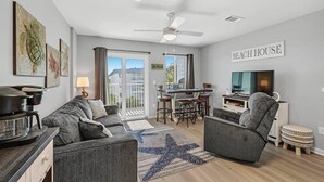 A modern living room with a grey sofa, a starfish rug, a dining area with a table and chairs, a ceiling fan, and a TV. - Tucked Away at Sea!!! (Gulf Shores)