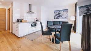 A modern kitchen with white cabinets, a built-in oven, and a stainless steel range hood. There is a dining area with a table and chairs, and a large window with curtains. - FP Appartements - Maisi 4, mountain view & summercard (Kaprun)