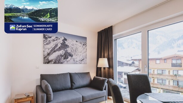 A modern hotel room with a gray sofa, a glass coffee table, and a large window offering a mountain view. - FP Appartements - Maisi 4, mountain view & summercard (Kaprun)