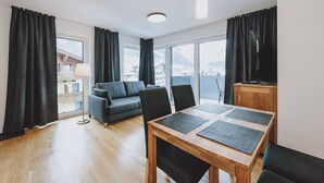 A modern living room with a wooden dining table, black chairs, a grey sofa, and a TV on a wooden cabinet. Large windows with black curtains offer a view of a mountainous landscape. - FP Appartements - Kitz 5, balcony & summercard (Kaprun)