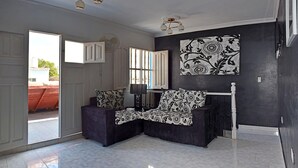 Ein modernes Wohnzimmer mit einer dunkelgrauen Sofagarnitur, einem großen Fenster mit Jalousien, einem Deckenventilator und einem Wandbild.
