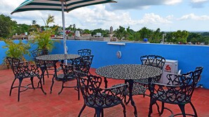 Eine Dachterrasse mit Tischen und Stühlen im Freien, einem Streifen-Sonnenschirm und einer blauen Wand.