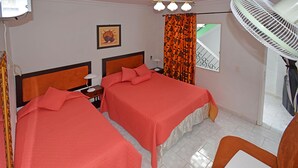 Ein Hotelzimmer mit zwei Betten, einem Schreibtisch, einem Stuhl, einem Ventilator, einem Bild an der Wand und einem Fenster mit Blick.