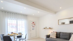 Un salon moderne avec un canapé gris, une table à manger où sont disposés des aliments pour le petit-déjeuner, et une grande fenêtre ornée de rideaux légers.