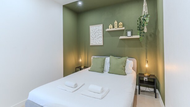 Un lit bien fait, avec du linge de lit blanc et des coussins verts, une lampe de chevet, un support à plantes en macramé fixé au mur et un tableau d’art abstrait encadré.