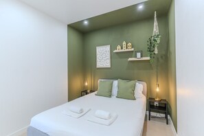 Een net opgemaakt bed met witte lakens en groene kussens, een bedlampje, een wandhanger van macramé voor een plant en een lijst met abstracte kunst.