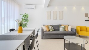 A modern living room with a grey sofa, yellow armchair, and a glass coffee table. There is a dining table with chairs and a yellow vase with green plants.