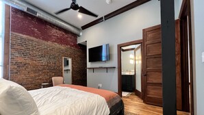 A bedroom with a brick wall, a bed with white bedding, a ceiling fan, a TV mounted on the wall, and a wooden door. - 1882 Lofts - 301-Downtown 2nd & Church (Nashville)