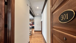 A hallway with a wooden floor, a number plate on a door, and a kitchen area in the background. - 1882 Lofts - 301-Downtown 2nd & Church (Nashville)