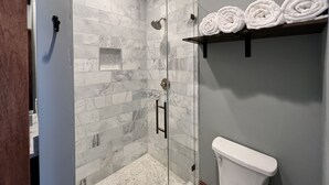 A bathroom with a glass-enclosed shower, marble tiles, a toilet, and a shelf with rolled towels. - 1882 Lofts - 301-Downtown 2nd & Church (Nashville)