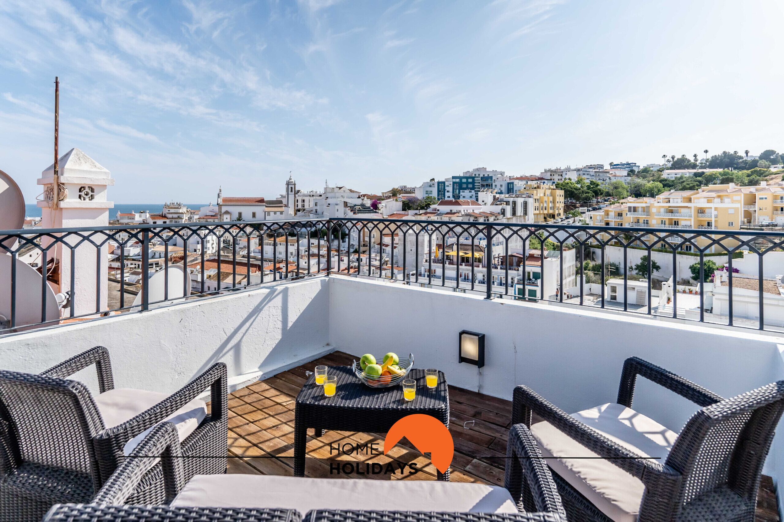 A rooftop terrace with wicker furniture, a table with fruit and drinks, and a cityscape view.