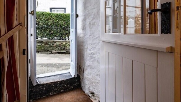 A narrow hallway with a white door leading outside, a window with a view of a hedge and a building, and a wooden door frame. - St Samson (Port Isaac)