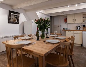 A dining area with a wooden table and chairs, a vase with flowers, and a kitchen in the background. - St Samson (Port Isaac)