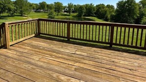 Eine Holzterrasse mit Geländer, die einen grünen Landschaftsbereich mit Bäumen und blauem Himmel überblickt.